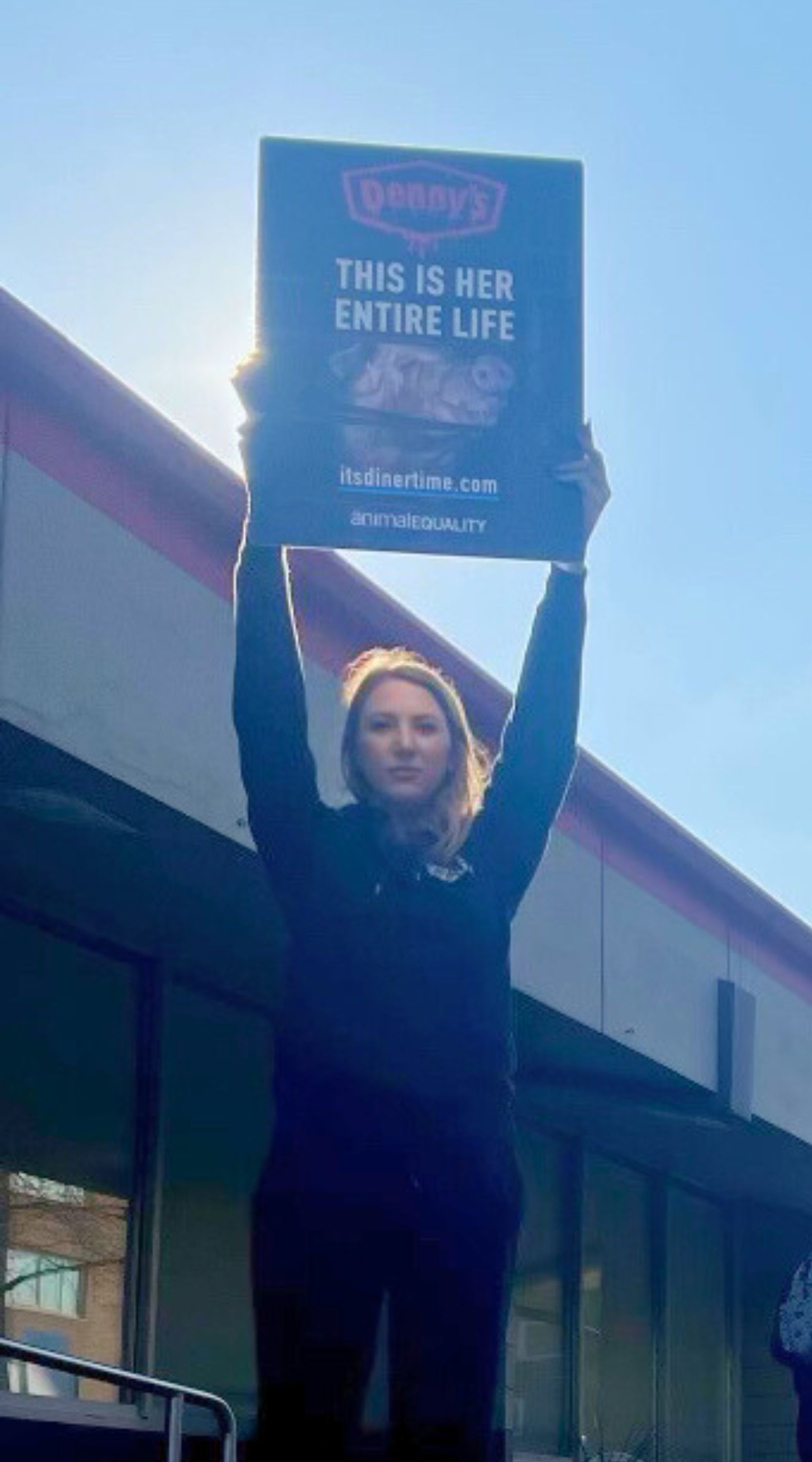 protestor standing with a sign that has a photo of a pig in a cage and the words "This is her entire life. itsdinertime.com"